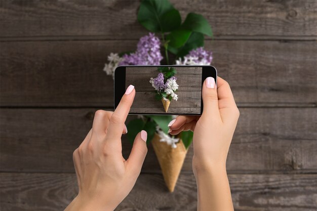 Photographer blogging workshop concept. Hand holding phone and taking photo of flowers. Lilac on wooden rustic background. Space for text. Hello spring