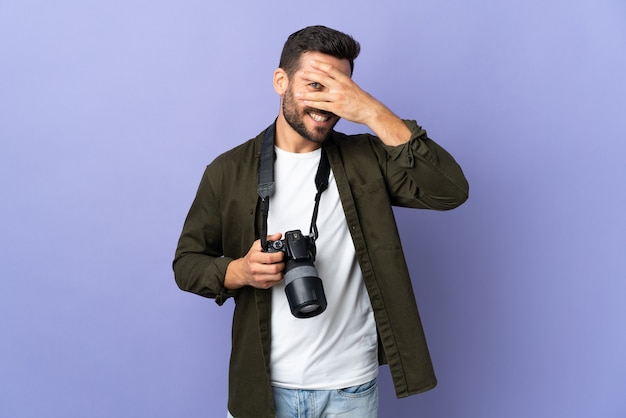 Photographer man over isolated purple background covering eyes by hands and smiling