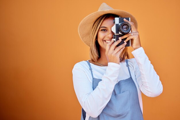 Photographer portrait and woman shooting a picture or photo with a retro camera isolated in an orange background Happy studio and female taking creative shots or fashion photographs as photography