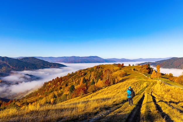 Photographer stands on ground grassy road making photo of autumn landscape with colorful trees and forestry mountains surrounded by fog at sunset