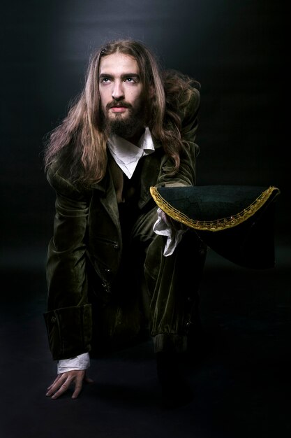 Photography medieval wary pirate man with long hair and beard, sat down on one knee looking away