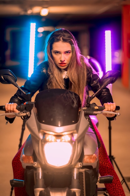 Photography with blue and pink neons on a motorcycle. Portrait of a young pretty blond Caucasian woman in a black leather jacket