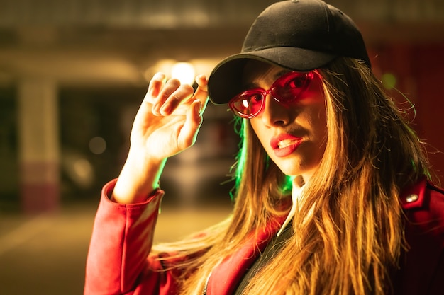 Fotografia con neon rossi e verdi in un parcheggio. ritratto di una giovane donna bionda caucasica in un vestito rosso, occhiali da sole e un berretto nero