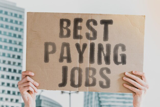 The phrase best paying jobs on a banner in men\'s hand with\
blurred background online wage profit make salary get gain earn\
vision skill paying top office treat high great pay