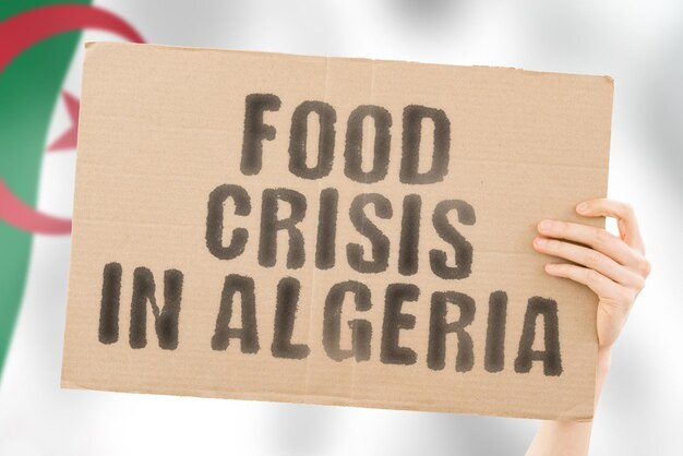 The phrase food crisis in algeria is on a banner in men\'s hands\
with a blurred algerian flag in the background crisis finance life\
nutrition bread disaster collapse social issue