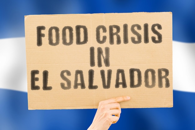 The phrase food crisis in el salvador is on a banner in men\'s\
hands with a blurred el salvador\'s flag in the background crisis\
finance life nutrition bread disaster collapse social issue