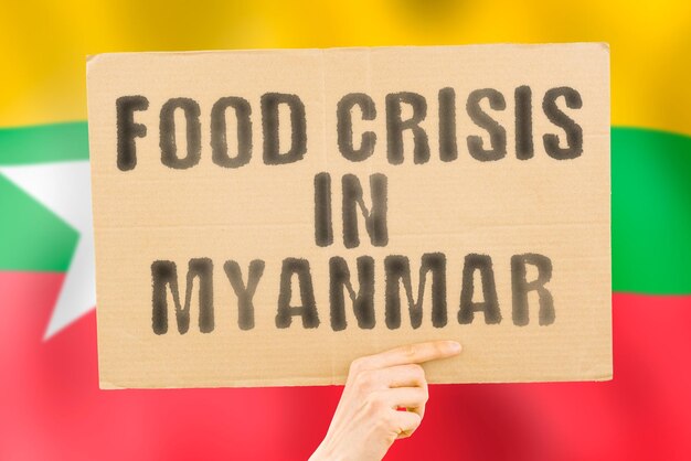 The phrase food crisis in maynmar is on a banner in men\'s hands\
with a blurred myanmarese flag in the background crisis finance\
life nutrition bread disaster collapse social issue