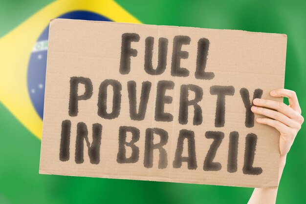 The phrase " Fuel poverty in Brazil " on a banner in men's hand with blurred Brazilian flag on the background. Economic. Expensive. Depressed. Gasoline. Fossil. Problem. Supply. Social issues