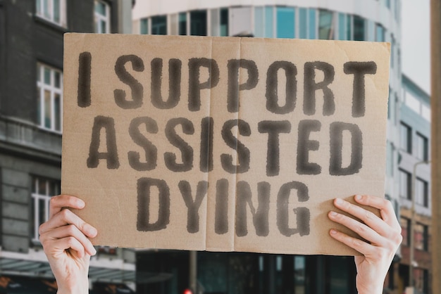 The phrase I support assisted dying is on a banner in men's hands with blurred background Treatment Assisted Health Kill Cancer Decision Die Lawyer Legal Option Pharmacy Sick End