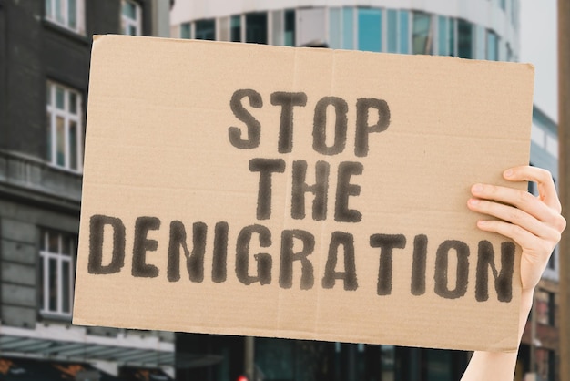 The phrase " Stop the denigration " on a banner in men's hand with blurred background. Harassment. Cruelty. Distrust. Cyberbulling. Victim. Separation. Suffer. Suffering. Trauma