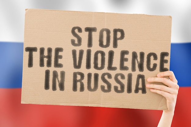 The phrase stop the violence in russia is on a banner in men\'s\
hands with a blurred russian flag in the background sad rights\
security social stress combat hate cruelty furious hitting