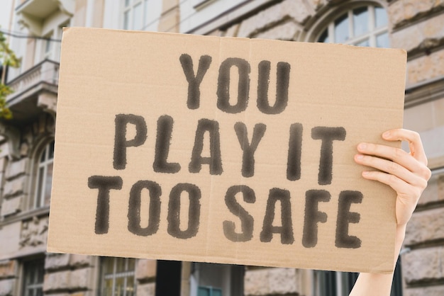 The phrase You play it too safe is on a banner in men's hands with blurred background Ride Risk Safe Safety Scare Scary Panic Person Phobia Play Power Problem