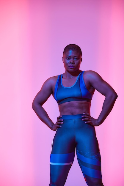 Photo physical activity full length vertical portrait of body positive young black female in sports top bra and leggings posing at studio looking at camera isolated on neon pink studio background banner