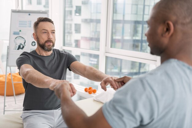 Physical exercise. Nice professional smart doctor holding his patients hands while doing a physical exercise with him