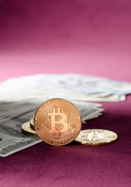 Physical Gold Bitcoin Coin against dollar bills on a purple background.