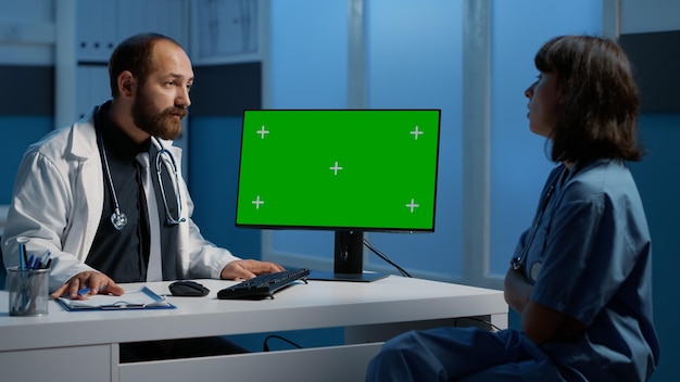 Physician and assistant discussing patient report planning\
health care treatment during checkup visit. doctor pointing at\
green screen computer template while working after hours in\
hospital office