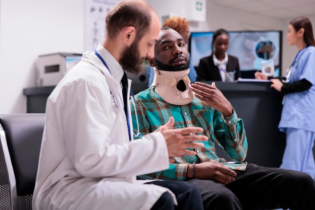 Physician consulting hurt person with neck collar foam, sitting in waiting room at reception lobby. Doctor helping injured man wearing cervical brace for support, checkup in waiting area.