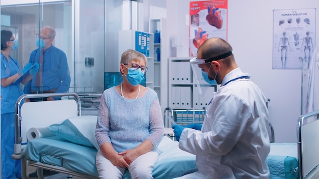 COVID-19 전염병 의료 글로벌 위기 동안 현대 개인 클리닉의 병원 침대에 앉아 있는 은퇴한 노부인의 폐 진단에 대해 논의하는 의사. coro에서 보호 장비에 의사