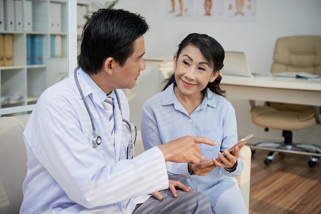 Photo physician explaining senior woman how to use telemedicine application on her smartphone for communication with doctor and checking her medical tests results