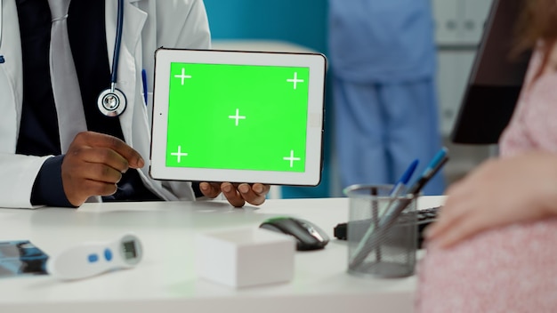 Physician and future mother looking at horizontal greenscreen on digital tablet. Male medic holding blank chroma key template with isolated copyspace and mockup background. Close up.