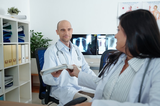 Physician giving folder with documents to her smiling colleague when they are working with papers in medical office