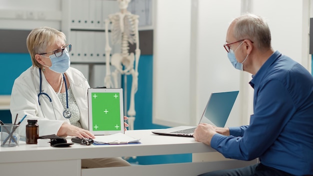 Physician showing digital tablet with greenscreen to old man in medical office during coronavirus pandemic. Blank isolated copyspace and mockup template with chroma key background.