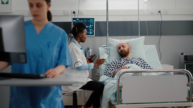 Physician with dark skin explaining pills treatment against pain during medical examination in hospital ward. Sick man patient with oxygen tube sitting in bed while doctor typing health expertise