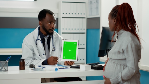 Physician and woman with baby bump looking at greenscreen on digital tablet in medical office. Doctor and patient analyzing isolated chroma key template with blank mockup copyspace.