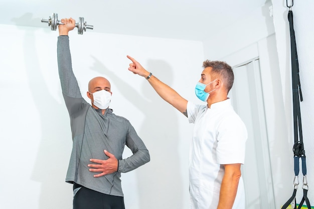 Physiotherapist with face mask and a patient doing exercises\
with a dumbbell physiotherapy with protective measures for the\
coronavirus pandemic covid19 osteopathy sports quiromassage