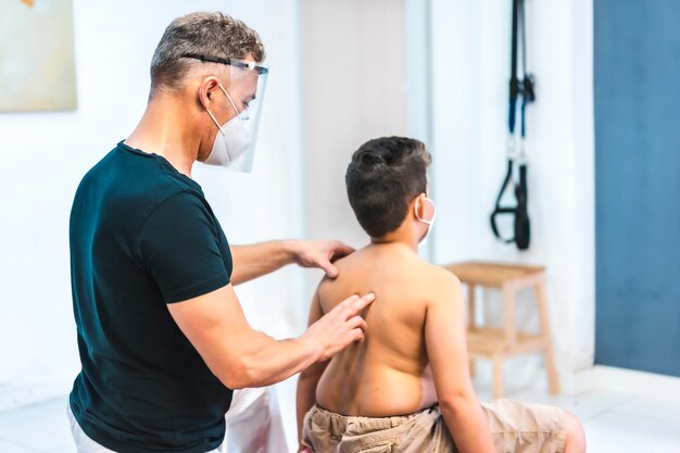 Physiotherapist with mask and protective screen doing a massage to a child. Reopening with security measures of physiotherapists in the Covid-19 pandemic. Osteopathy, therapeutic chiromassage