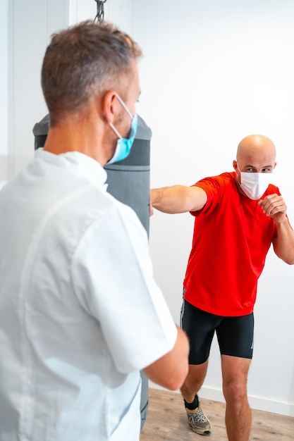 Fisioterapista con mascherina insieme al paziente sul sacco da boxe fisioterapia con misure di protezione per la pandemia di coronavirus covid19 osteopatia quiromassaggio sportivo