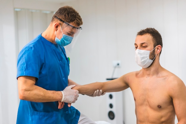 Физиотерапевт с экраном и маской дает массаж запястья пациенту с маской. Физиотерапия с защитными мерами при пандемии коронавируса, COVID-19. Остеопатия