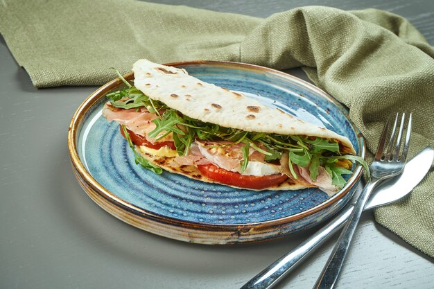 Piadina - klassiek Italiaans tortilla-brood met ham, rucola, mozzarella en tomaten op een blauw bord op houten tafel. Snelle sandwich