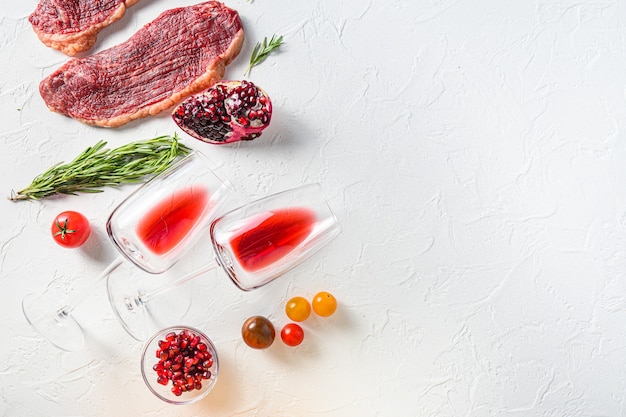 Органические стейки из говядины Picanha с розмарином, перцем, гранатом, около красного вина в очках и бутылке на белом текстурированном фоне, вид сверху с пространством для текста.