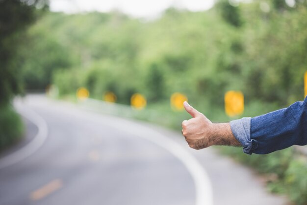 Foto prendimi. uomo che fa l'autostop sul ciglio della strada. l'uomo prova a fermare l'auto con il pollice in su. fare l'autostop è uno dei modi più economici di viaggiare.