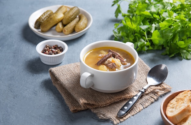 Pickle (Russian soup). Pearl soup with pickles and potatoes in a white plate on a blue background with bread and fresh  herbs. Copy space image