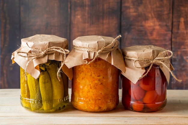 Pickled farm vegetables in glass jars on a wooden background.