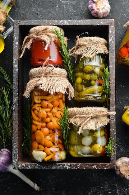 Pickled vegetables and mushrooms in glass jars in Wooden box on black stone background Top view