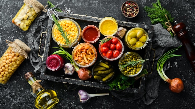 Pickled vegetables and mushrooms in glass jars in Wooden box on black stone background Top view