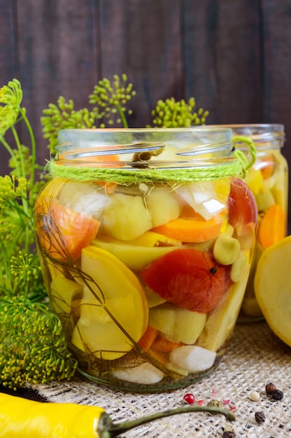 Photo pickles: vegetable assortment (zucchini, pepper, carrots, tomato, green peas) in glass jars on a dark wooden.