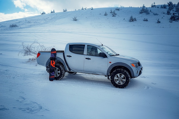 道路上のピックアップトラック、雪山の下の美しい冬の道。雪の深さを測る光沢のあるシルバーのトラック。どんな天候でも移動の自由のアイデアとコンセプト