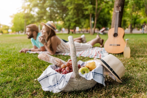 Picnic basket in focus, young couple on the grass in the park relaxing with guitar 