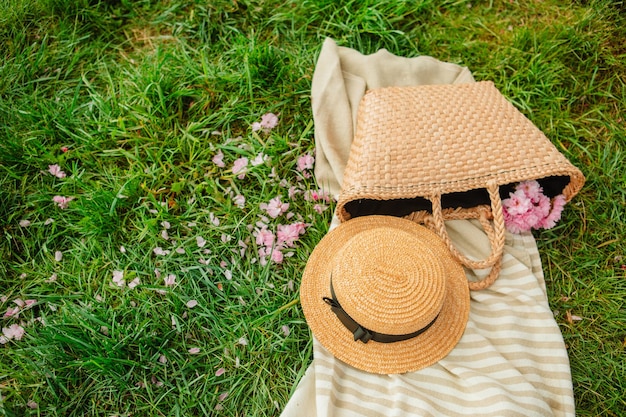 Picnic blanket with straw hat and bag on green grass covered with pink sakura flowers copy space