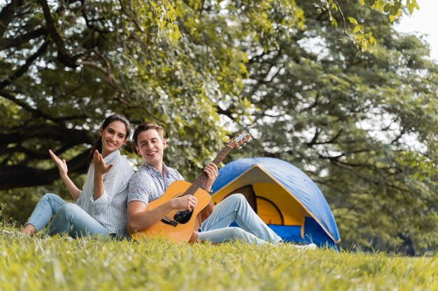 ピクニックとキャンプの時間。若いカップルがピクニックや公園でのキャンプでギターを楽しんでいます。愛と優しさ、彼のガールフレンド、ライフスタイルコンセプトにギターを弾くロマンチックな男