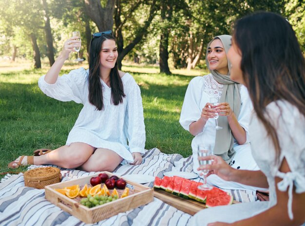 夏の屋外の公園でピクニック シャンパンと女性の乾杯 週末の自然の中でのフード ドリンクと幸せな友人のグループ 多様性の友情と芝生でのパーティーは、夏にリラックスします