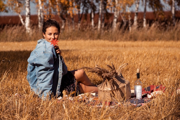 Picnic in the fresh air: a young woman in a denim jacket and dress is holding a red leaf and enjoying nature, sitting on a plaid with a picnic basket, apples, wine. 