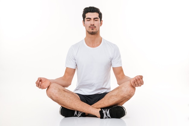 Picture of attractive sportsman in gym sitting on floor over white wall. Eyes closed. Yoga concept.