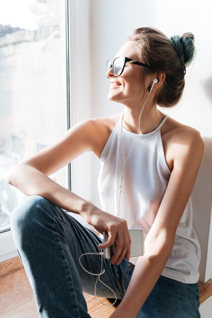 音楽を聴いている家の窓の近くに座っている眼鏡をかけている魅力的な若い女性の写真。ウィンドウを見てください。