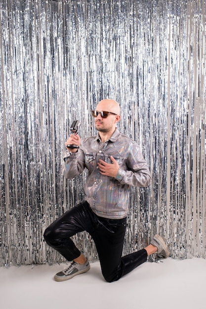 Picture of beautiful caucasian man without hair with black sunglasses in a shiny shirt, black leather pants and grey sneakers sings in a small silver microphone