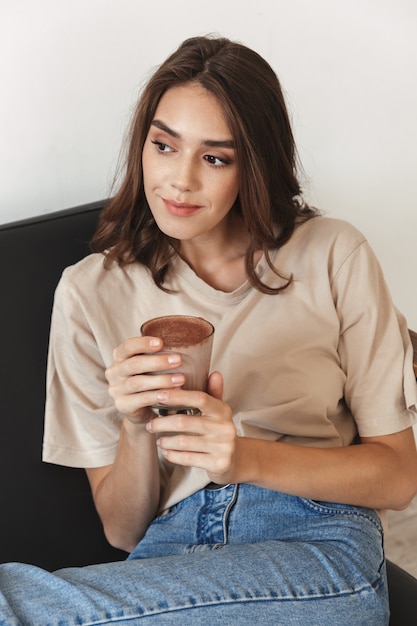 自宅の屋内でコーヒーやお茶のカップを保持しているソファで美しい楽観的な若い驚くべき女性の写真。